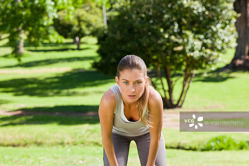 穿着运动服的女性在阳光明媚的草地公园中，双手放在膝盖上，向前倾身暂停。图片素材