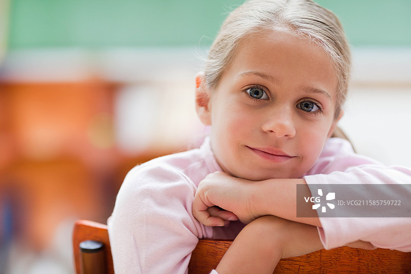 穿着粉色上衣的女孩靠在木椅上，凝视着教室里的黑板。图片素材