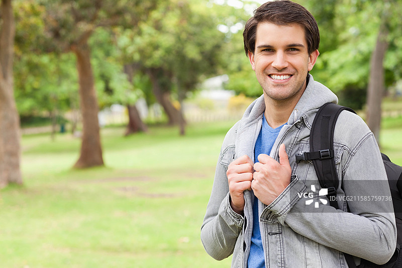 公园里一个穿着灰色夹克、蓝色衬衫、背着黑色背包的微笑男子，留白空间。图片素材