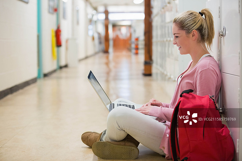 青少年学生坐在学校走廊里，使用银色笔记本电脑学习，旁边放着红色背包，留有空白空间。图片素材