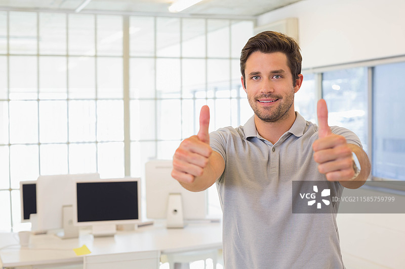 男子在明亮的现代办公室中竖起两个大拇指，办公室内有桌子、显示器和玻璃隔断。图片素材