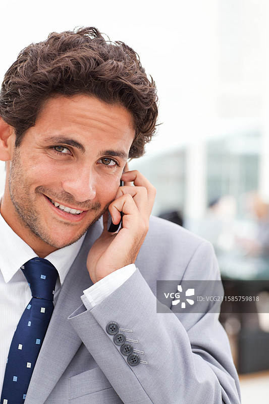 商人在现代办公室中微笑着打电话，手持智能手机，周围有玻璃隔断。图片素材
