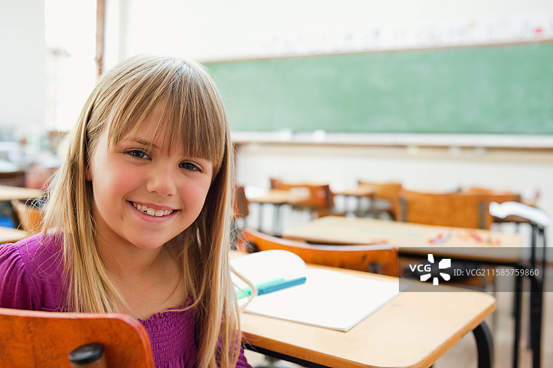 微笑的女童坐在教室的木制书桌前，手里拿着笔和笔记本，留有空白空间。图片素材