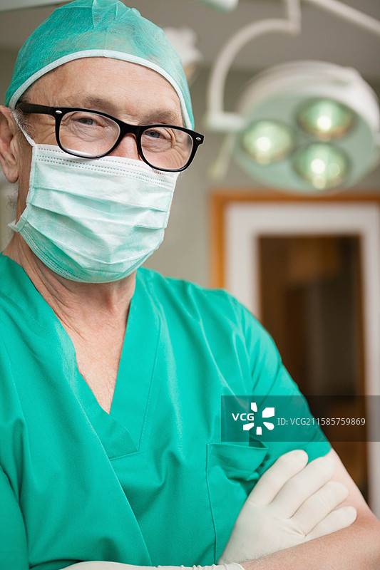资深外科医生穿着绿色手术服、帽子、口罩和手套，站在手术室的手术灯下。图片素材