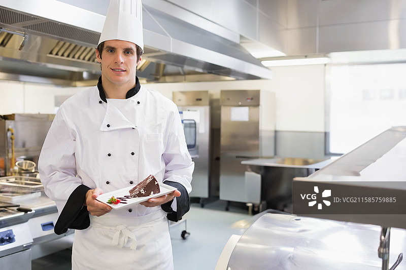 穿着制服的男厨师在厨房里靠近烤箱，手里拿着一块装饰有草莓的巧克力蛋糕。图片素材