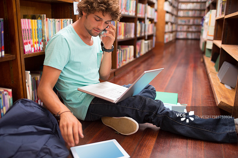 图书馆里的青少年学生使用笔记本电脑，手持智能手机、平板电脑，背着背包，旁边有笔记本，留有空白空间。图片素材