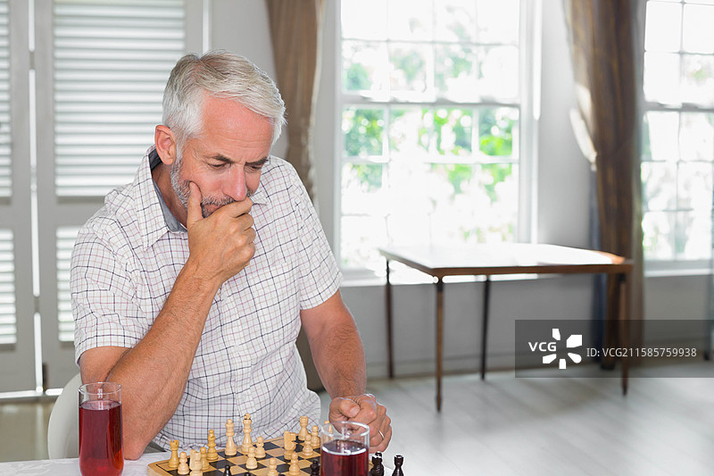 老年男子坐在家中的桌子旁，手握一杯红酒，思考着棋盘上的下一步。图片素材