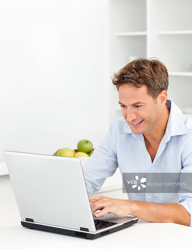 中年男子穿着衬衫，坐在厨房桌子前微笑着用笔记本电脑打字，桌上有一碗苹果。图片素材
