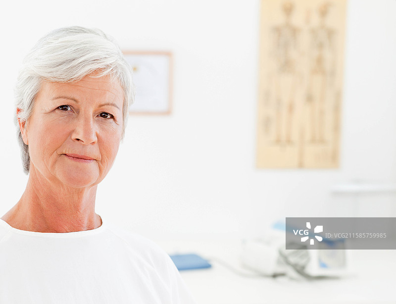 一位老年女性站在考试室内，旁边有骨骼海报、证书和血压监测仪，留有空白空间。图片素材