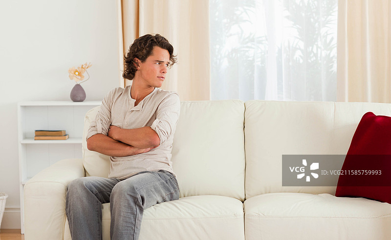 男人坐在米白色沙发上，目光向右凝视，家中有红色抱枕和干花花瓶，留白空间。图片素材