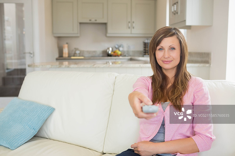 女人坐在沙发上，手握遥控器，旁边是靠垫和厨房台面的水果碗，留有空白空间。图片素材