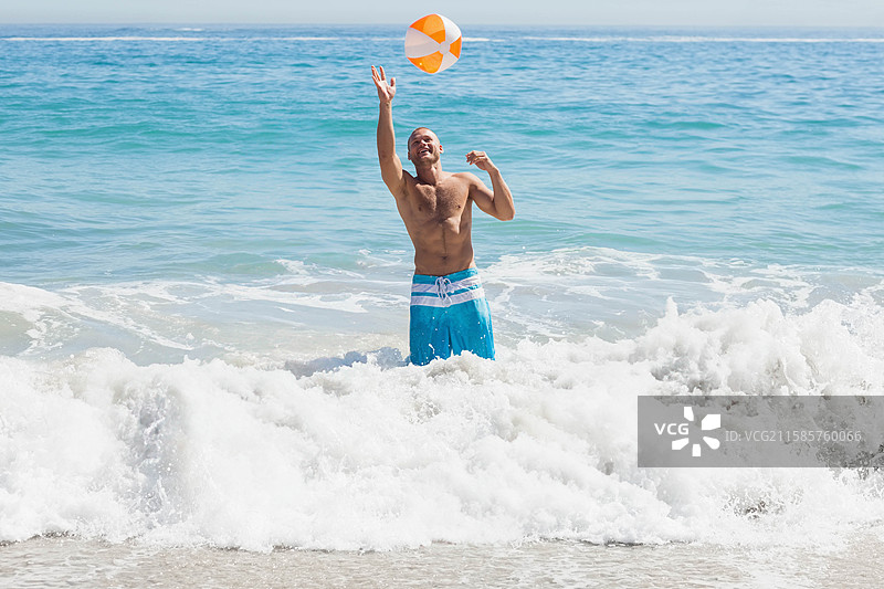 赤裸上身的男子穿着泳裤，站在海滩的波浪中，抛掷着一只橙白相间的沙滩球。图片素材