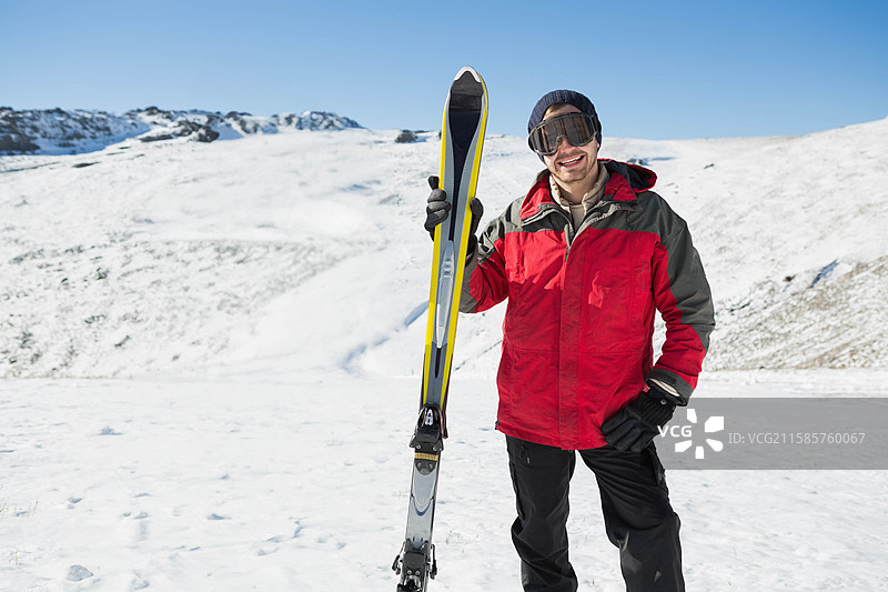 单身男性滑雪者站在山坡上，穿着滑雪夹克和手套，手里拿着滑雪板。图片素材