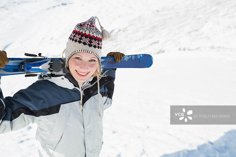站在雪覆盖的斜坡上的女性，穿着冬季外套，戴着针织帽，手持蓝色滑雪板，留有空白空间图片素材