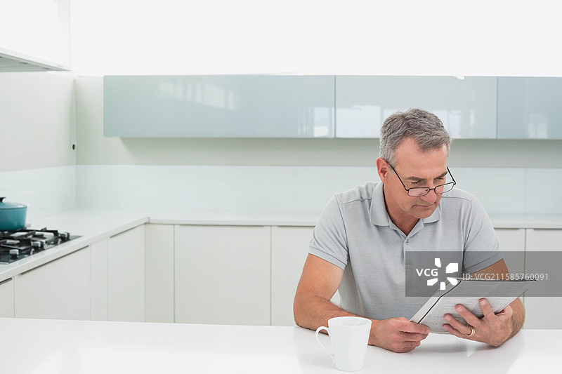 男人坐在厨房岛上阅读报纸，手边有咖啡杯和青绿色的壶，留有空白空间。图片素材