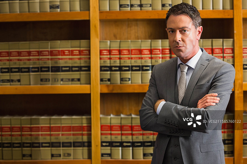 身穿灰色西装和领带的男子双臂交叉，站在法律图书馆的书架前，留有空白空间。图片素材