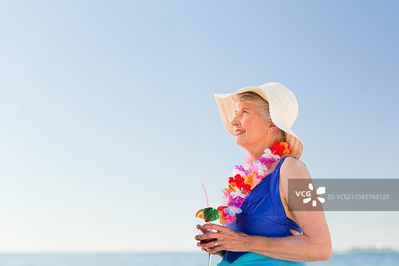 一位年长女性在海滩上手持花环和带伞的鸡尾酒杯，留有空白空间。图片素材