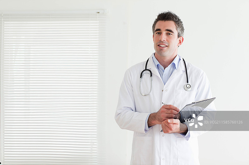 男医生在诊所的检查室里，靠窗帘写在夹板上的笔记，身边有听诊器和钢笔。图片素材