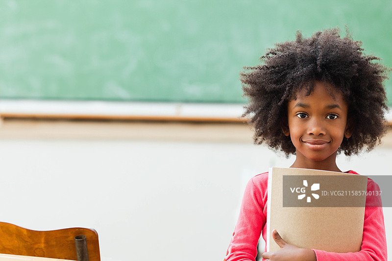 非裔美国女孩站在学校里，手里拿着棕色笔记本，旁边是绿色黑板，留有空白空间。图片素材