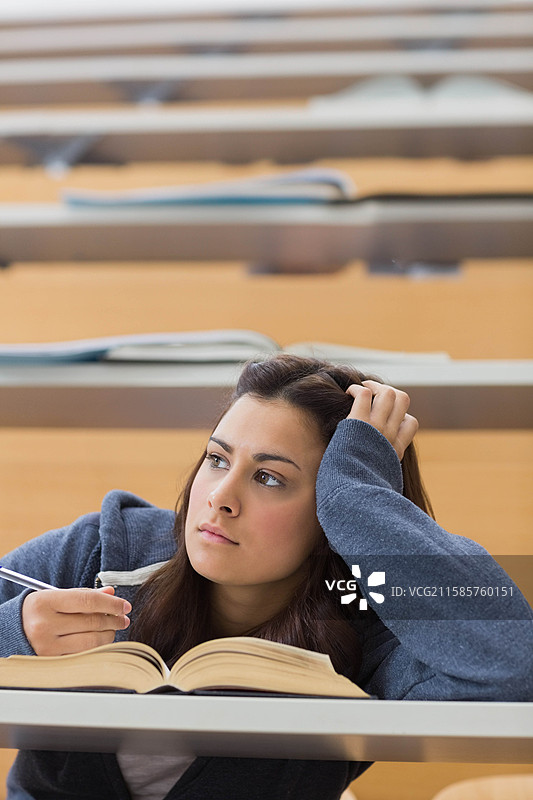 女学生在大学讲堂的分层木桌上拿着笔，正在学习打开的教科书。图片素材