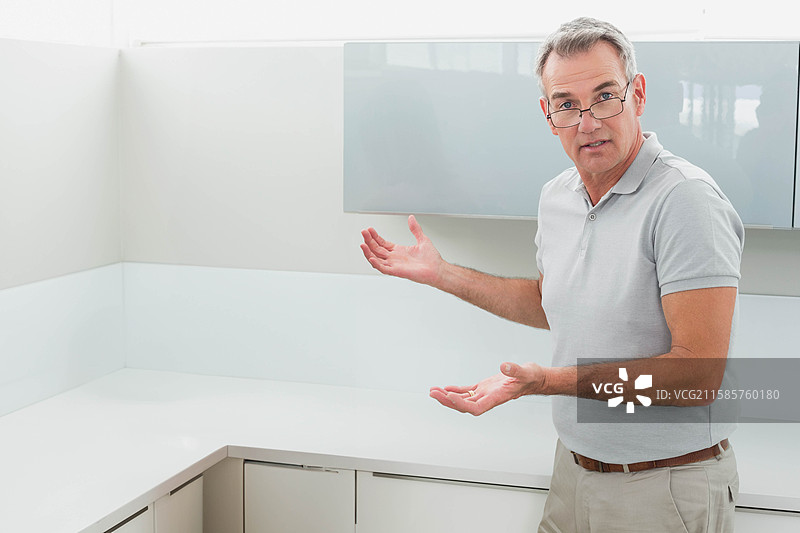 老年男子站在厨房里，展示白色台面和反光橱柜，留有空白空间图片素材