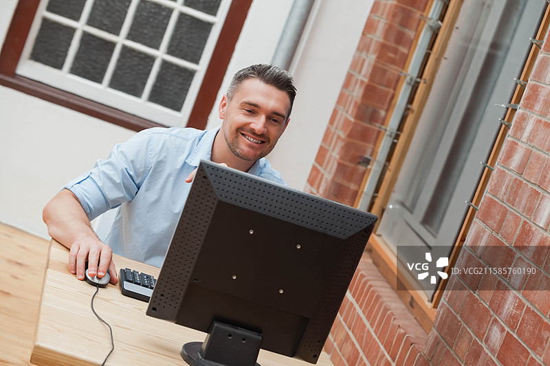 中年男子坐在木质办公桌前，微笑着使用台式显示器和有线鼠标。图片素材