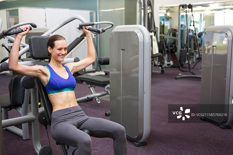 女性健身者穿着运动胸罩和紧身裤，在健身房使用肩部推举机，留有空白空间图片素材