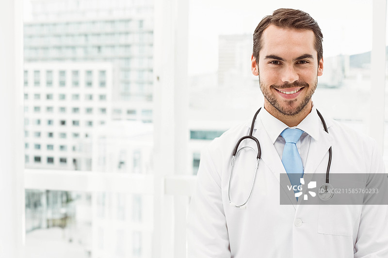 男医生穿着白大褂和听诊器，站在诊所的玻璃窗旁，留有空白空间。图片素材