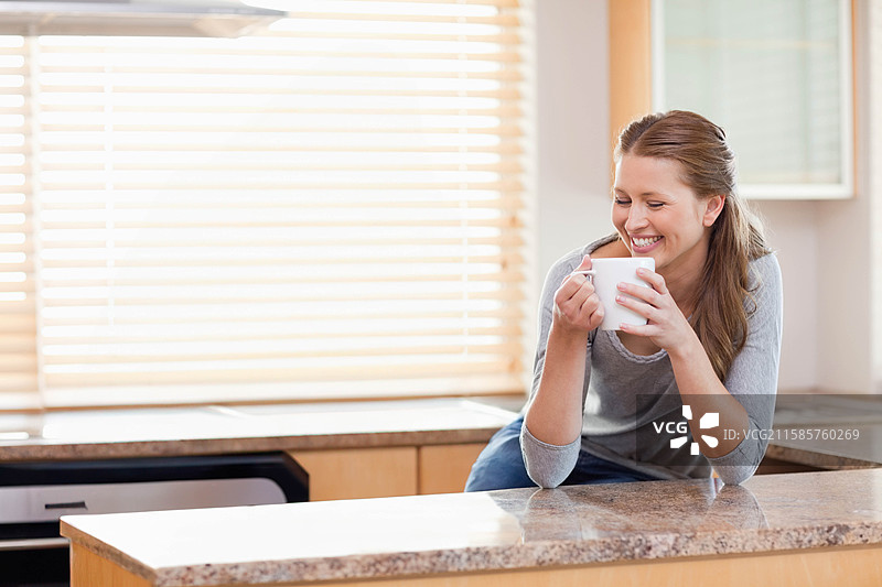 女人靠在花岗岩厨房岛上，手里拿着陶瓷杯，窗帘下有空白空间。图片素材