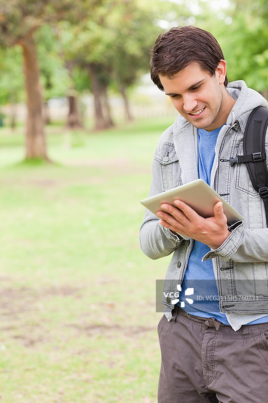在公园里站着的男人，手持平板电脑，微笑着，穿着夹克，背着背包，留有空白空间。图片素材