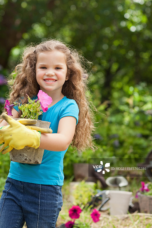 女孩在阳光明媚的后院花园里，戴着黄色手套，手里拿着一个装满粉色矮牵牛的混凝土花盆。图片素材