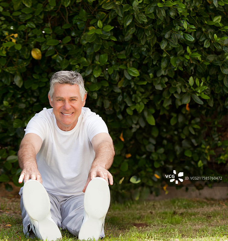 老年男子在后院花园的草地上向前伸展，试图触碰脚趾，穿着白色运动鞋。图片素材