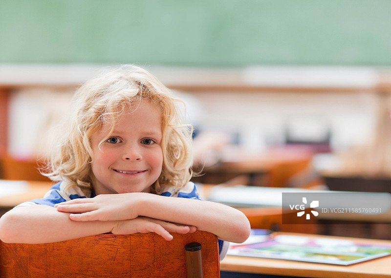 男孩靠在教室里的木椅上，背景是绿色黑板和五彩的练习册。图片素材