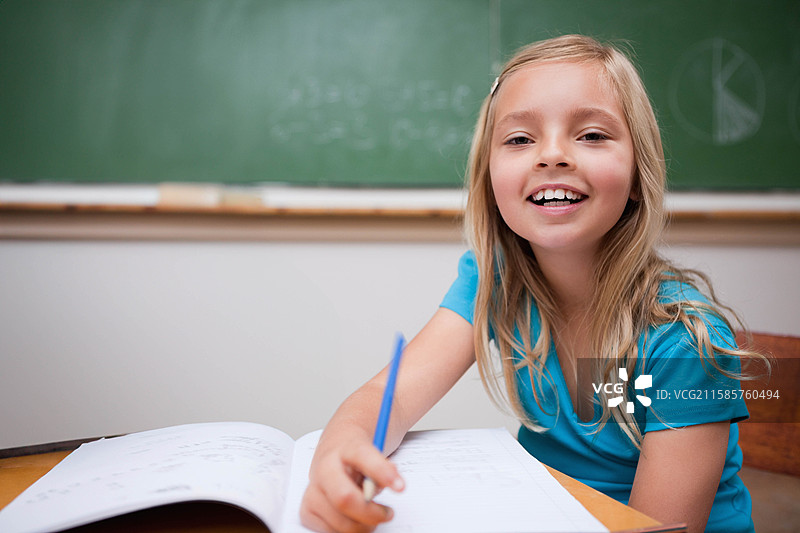女童坐在教室的桌子前，用笔在作业本上写字，背景是绿色黑板，留有空白空间。图片素材