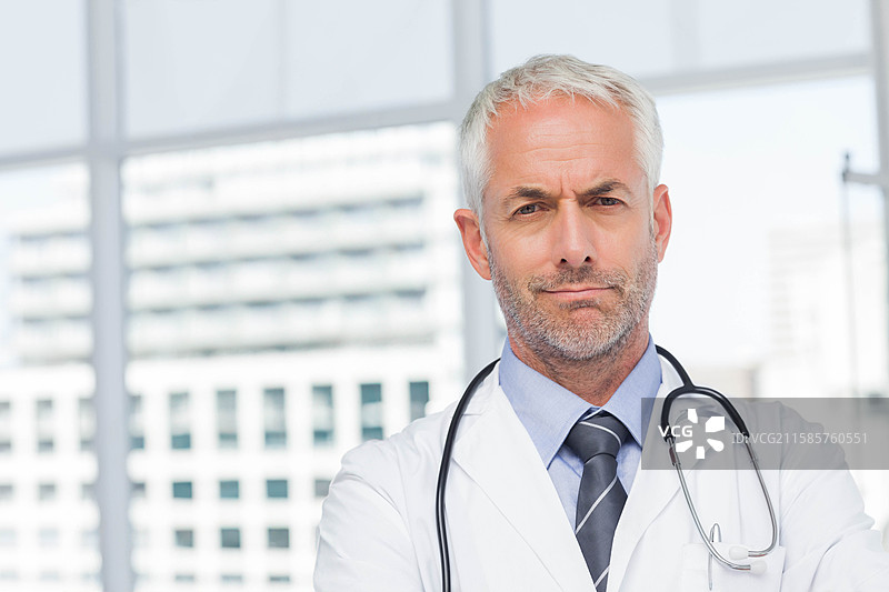 穿着白大褂和听诊器的老年男性医生站在诊所的窗边，留有空白空间图片素材