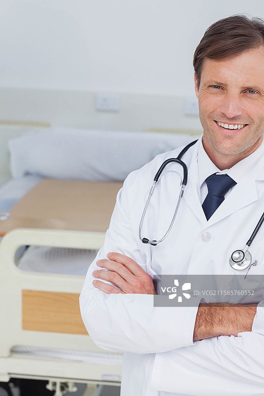 穿着听诊器的男性医生双臂交叉，站在医院病床旁，床上有床单和枕头，留有空白空间。图片素材