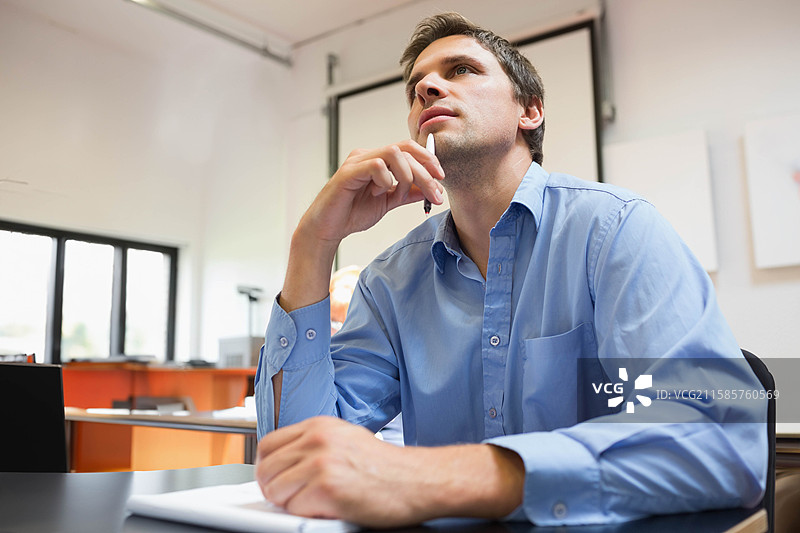 男子坐在会议室的桌子旁，手持笔靠在下巴上，注视着投影屏幕和笔记本。图片素材