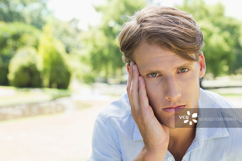 男人坐在绿树成荫的公园里，手托着脸深思，旁边有一个池塘，留有空白空间。图片素材