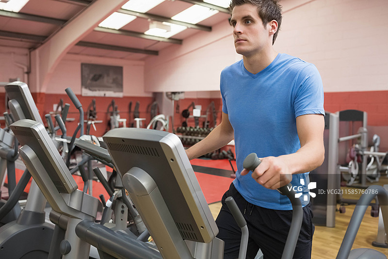 男子在商业健身房的椭圆机上锻炼，旁边有哑铃架和长椅，留白空间图片素材