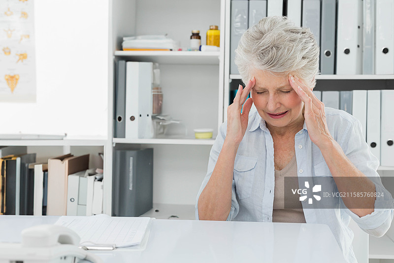 老年女性在诊所桌前用手按压太阳穴，旁边有电话和笔记本，留有空白空间图片素材