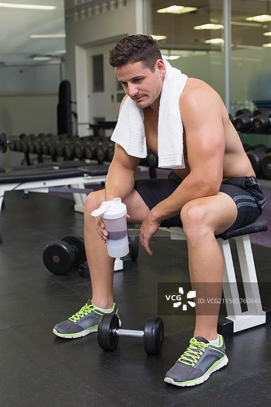 男性健身爱好者坐在健身房的举重凳上，手拿毛巾，正在用摇摇杯喝水。图片素材