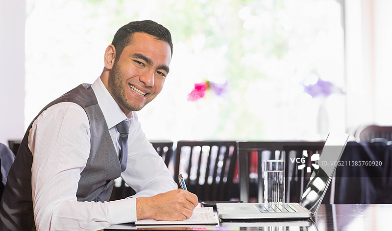 身穿商务服装的男子在咖啡馆桌子上做笔记，旁边有笔记本电脑和笔记本，留有空白空间图片素材