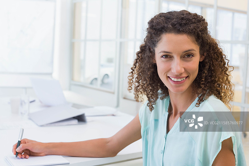 女人在办公桌前微笑着看向镜头，手持笔，旁边有笔记本和笔记本电脑，隔着玻璃隔断。图片素材