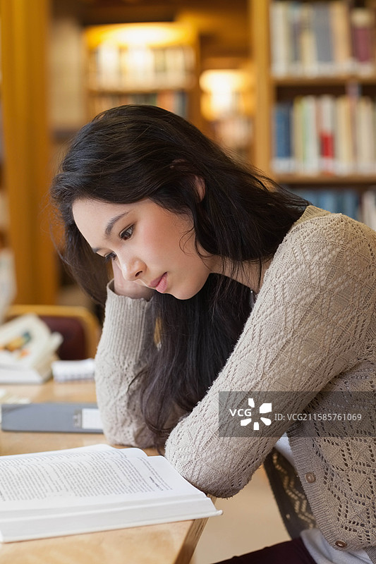 亚洲女性靠在手上，阅读放在木质图书馆学习桌上的打开教科书，旁边有笔记本和钢笔。图片素材