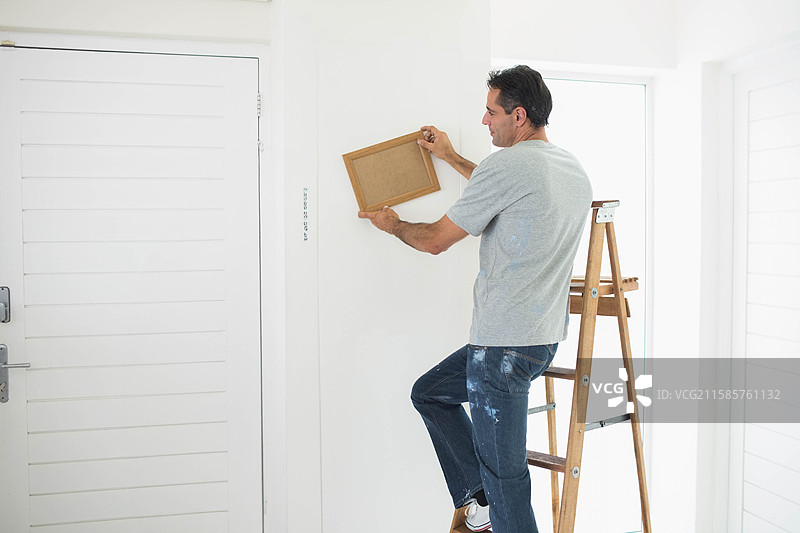 在白色走廊墙上用梯子将木质相框调整水平，靠近关闭的窗户。图片素材