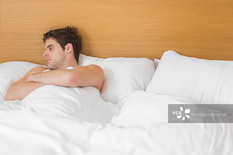 男子靠在木制床头板上，穿着无袖内衣，盖着被子，留有空白空间。图片素材