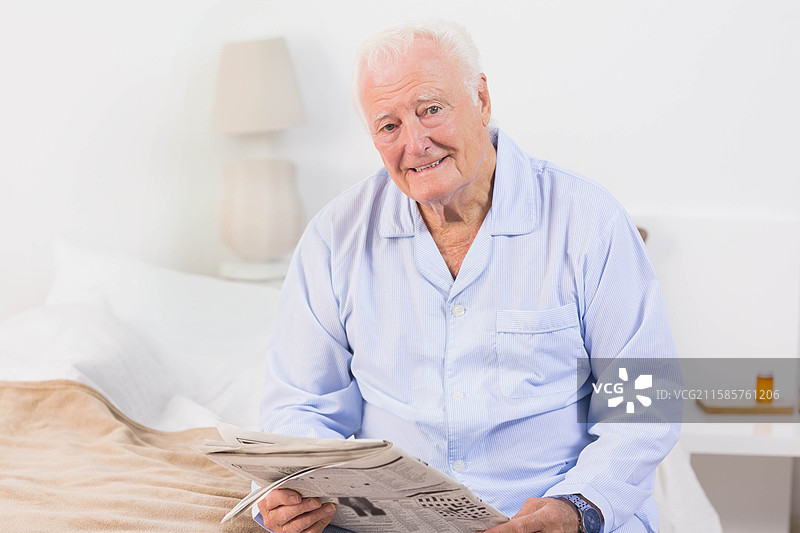 穿着睡衣的老年男子坐在卧室里，灯光下阅读报纸，旁边有药瓶。图片素材