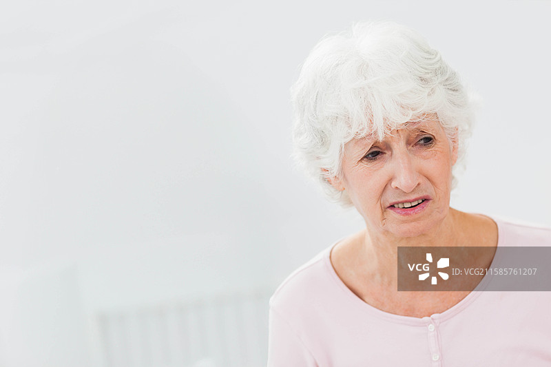 一位穿着浅粉色上衣的老年女性坐在明亮房间的壁挂式暖气旁，留有空白空间。图片素材