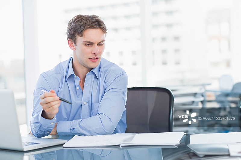 男性商务专业人士在现代办公室的玻璃桌上审阅文件，桌上有笔和笔记本电脑。图片素材