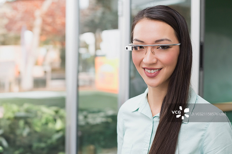 亚洲女性站在现代办公室大堂内，佩戴智能眼镜，靠近大型玻璃面板。图片素材